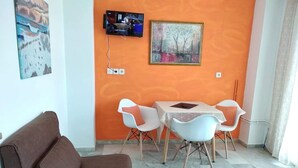 A living room with a brown sofa, a TV mounted on the wall, and a painting. There is a small table with chairs in front of the sofa. - Poseidon Apartments (Kefalonia)