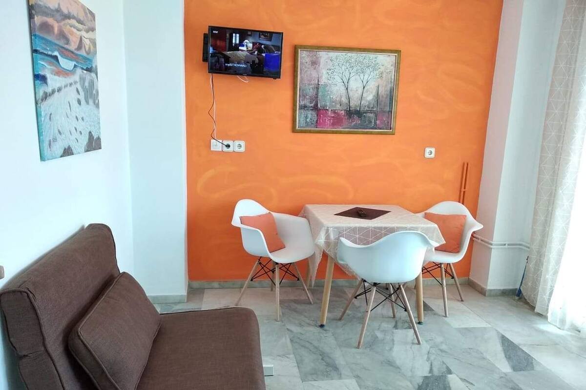 A living room with a brown sofa, a TV mounted on the wall, and a painting. There is a small table with chairs in front of the sofa.