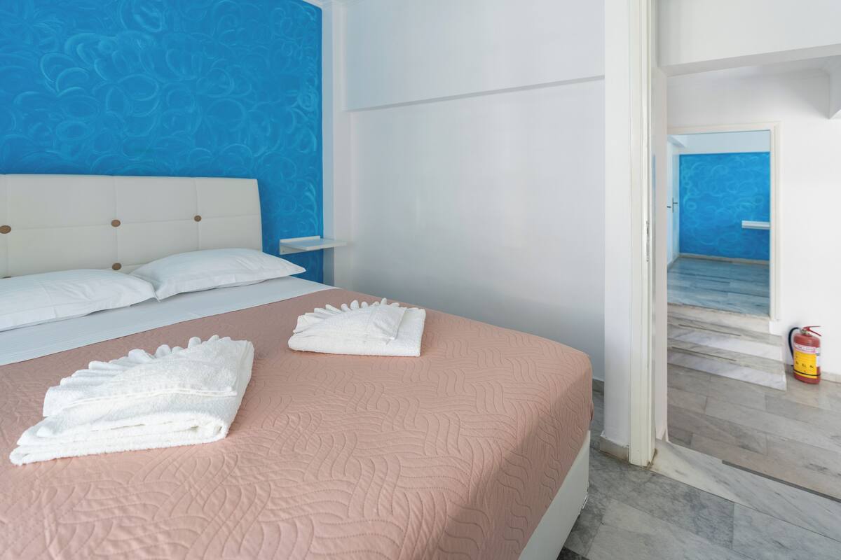 A neatly made bed with white linens and a pink quilt, a white headboard, and a blue patterned wall.