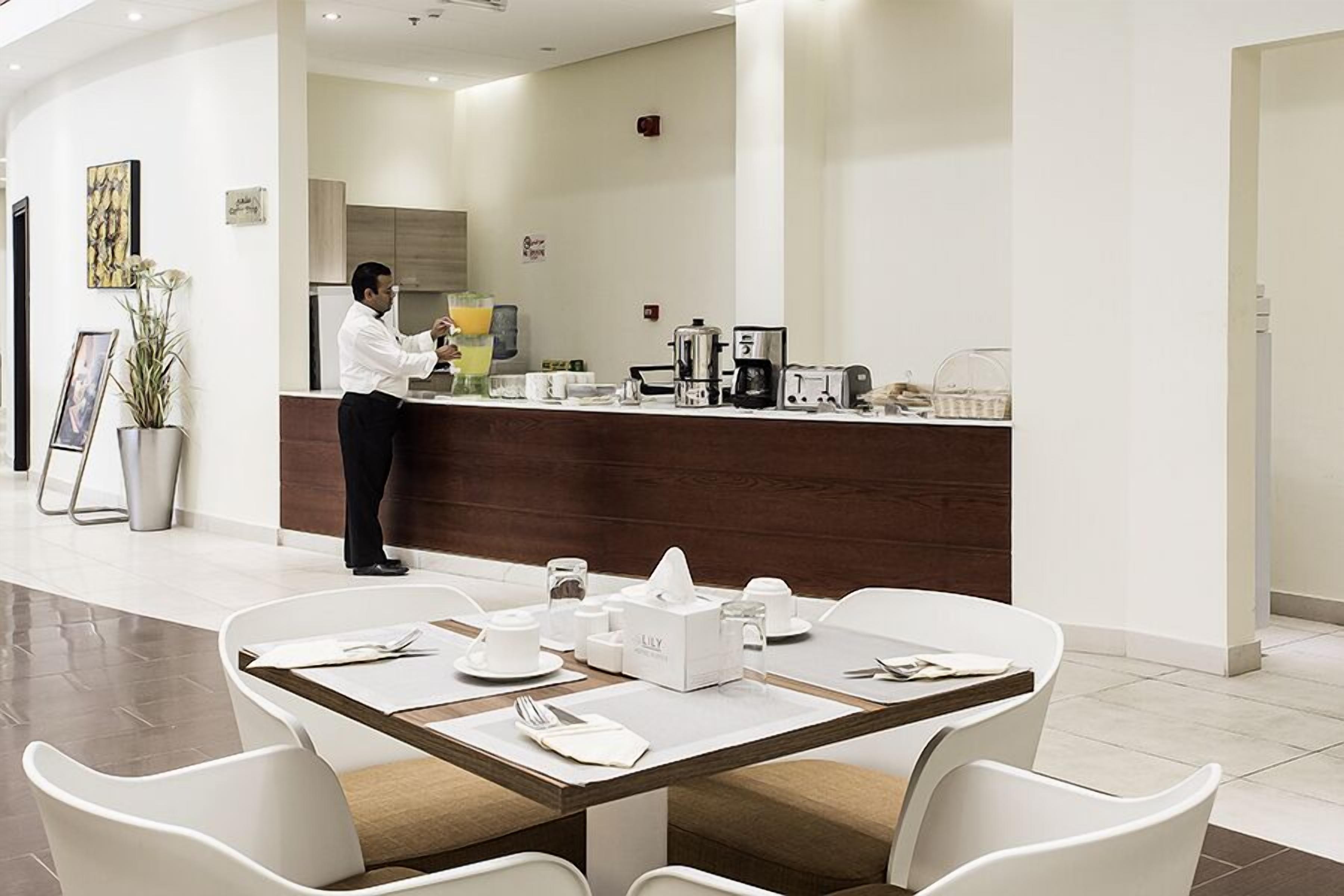 A modern hotel lobby with a reception desk, a coffee machine, and a dining area with white chairs and a wooden table.