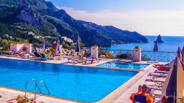 Un espace piscine doté de chaises longues et de parasols, offrant une vue sur une côte montagneuse.
