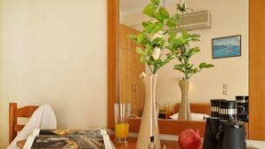 Une chambre d’hôtel avec une table en bois, un vase contenant des plantes vertes, une grenade, un verre de jus d’orange, un livre et des jumelles.
