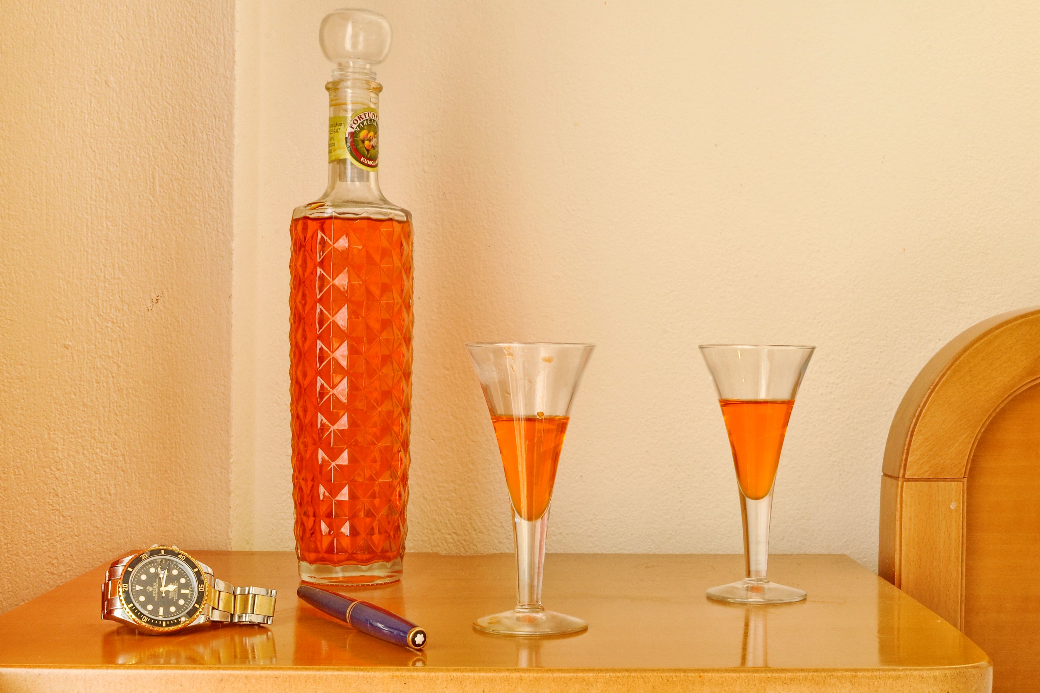 A bottle of liquor, two glasses, and a wristwatch on a wooden table.