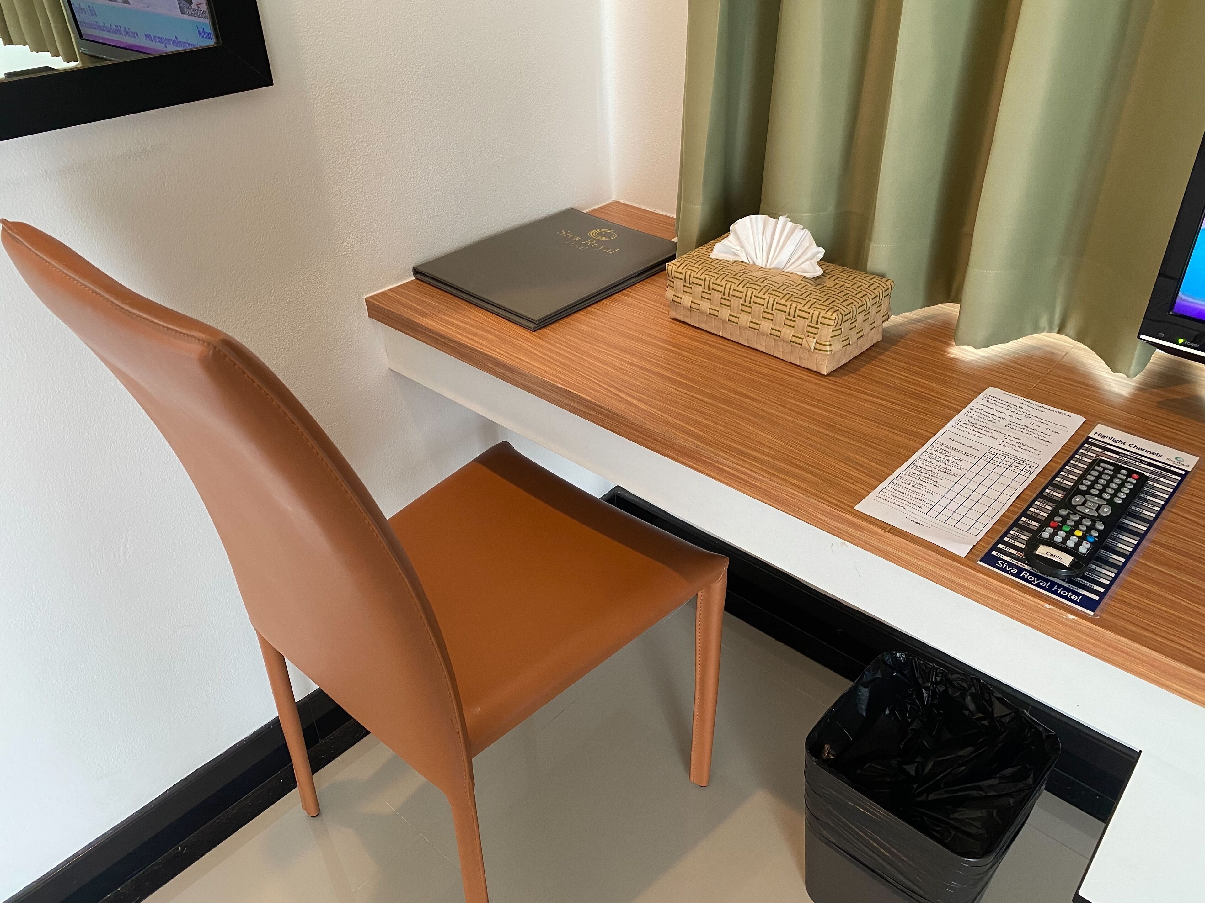 A hotel room with a wooden desk, a laptop, a tissue box, a remote control, and a trash bin.