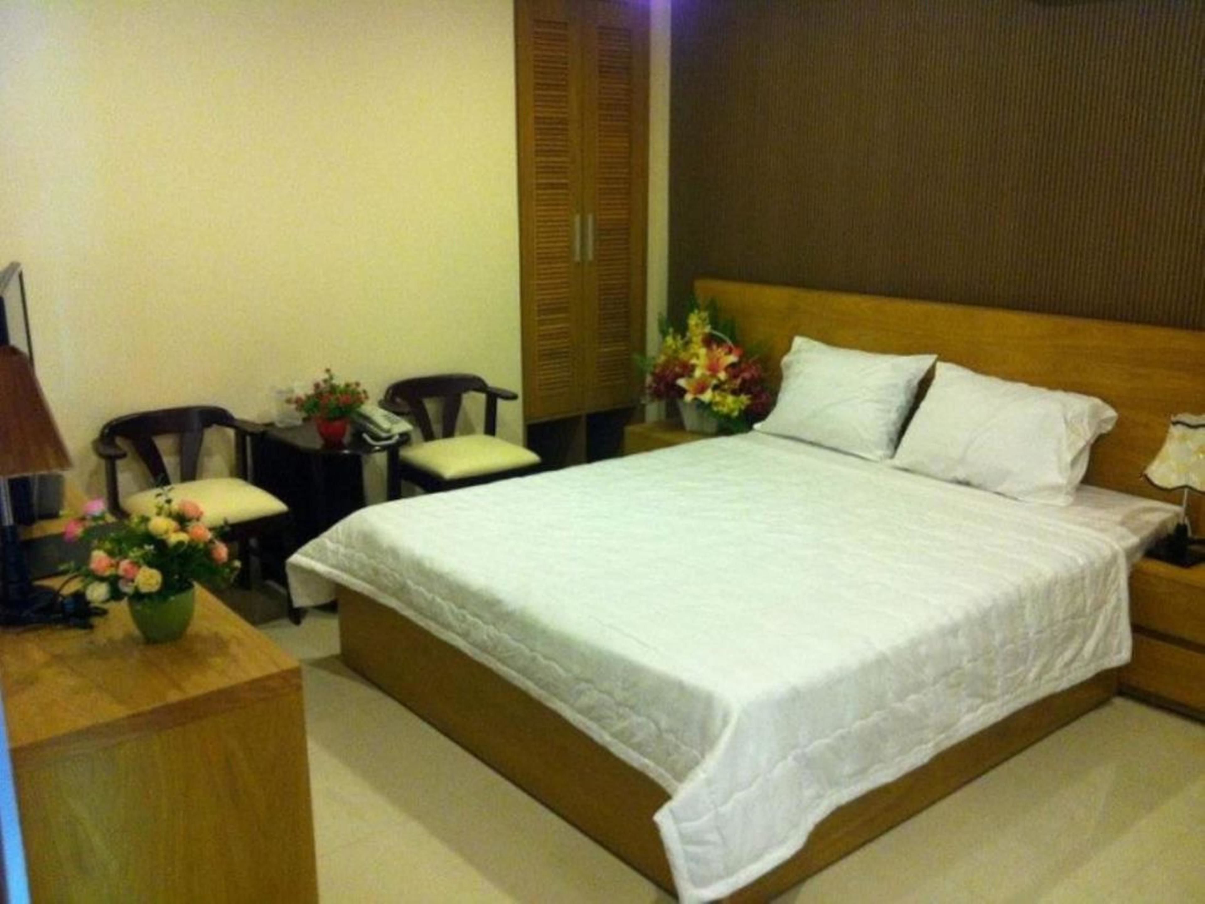 A neatly arranged hotel room with a large bed, a wooden nightstand, a desk with a telephone, two chairs, and a small table with flowers.