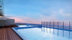 Una piscina infinita en la azotea con terraza de madera y sillas reclinables, con vistas a un paisaje urbano.