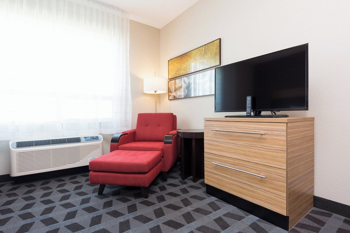 Une chambre d’hôtel avec un fauteuil rouge, une commode en bois, un téléviseur à écran plat et une fenêtre avec des stores.
