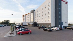 Un hotel de diseño moderno, con estacionamiento que muestra varios autos y un pequeño espacio ajardinado con un árbol.