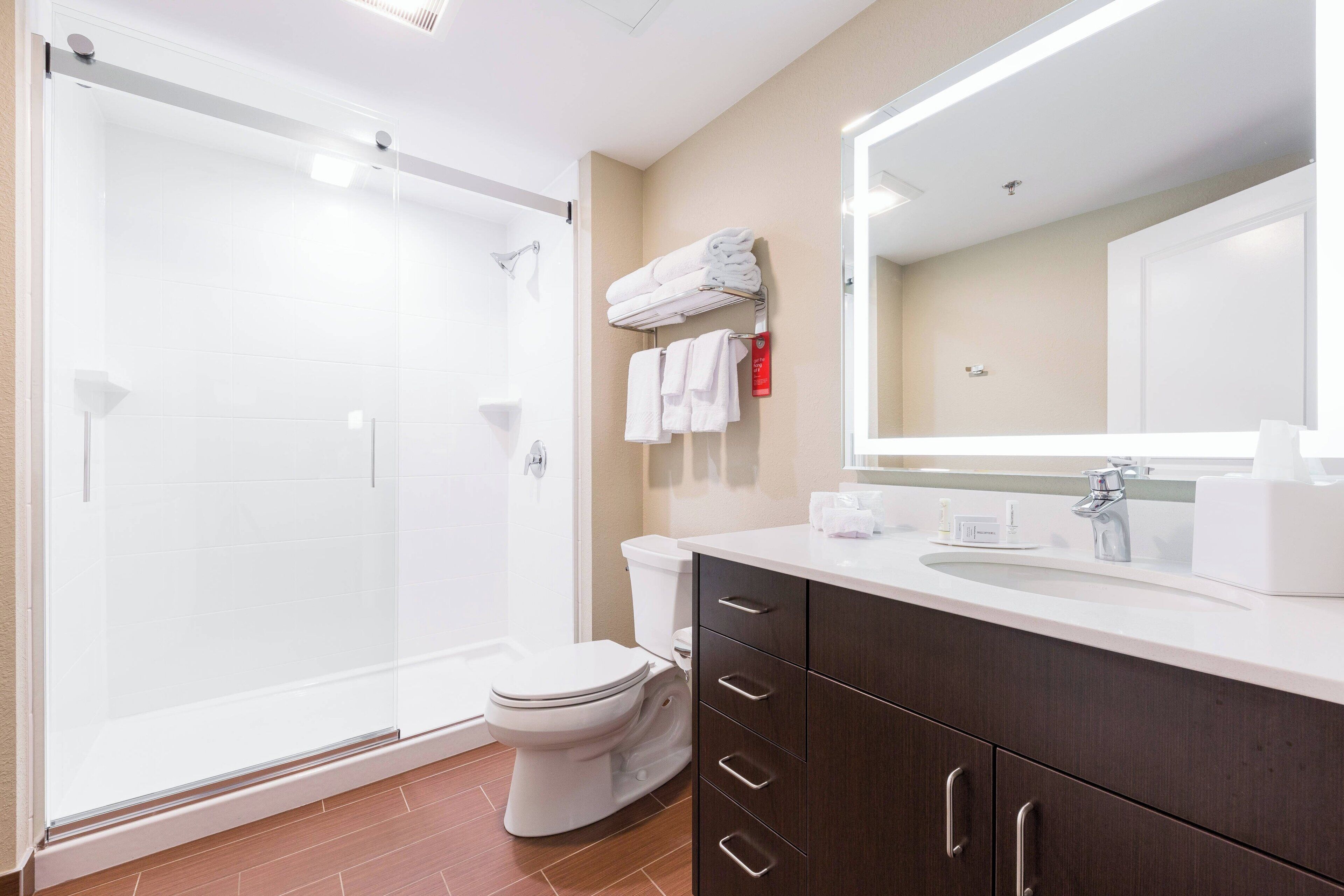 Un baño moderno con ducha acristalada, inodoro blanco, tocador con espejo y toallas ordenadas en un perchero.