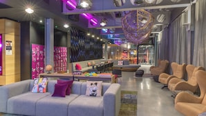 A modern hotel lobby with a grey sofa, beige armchairs, a dining area, and a large decorative hanging light.