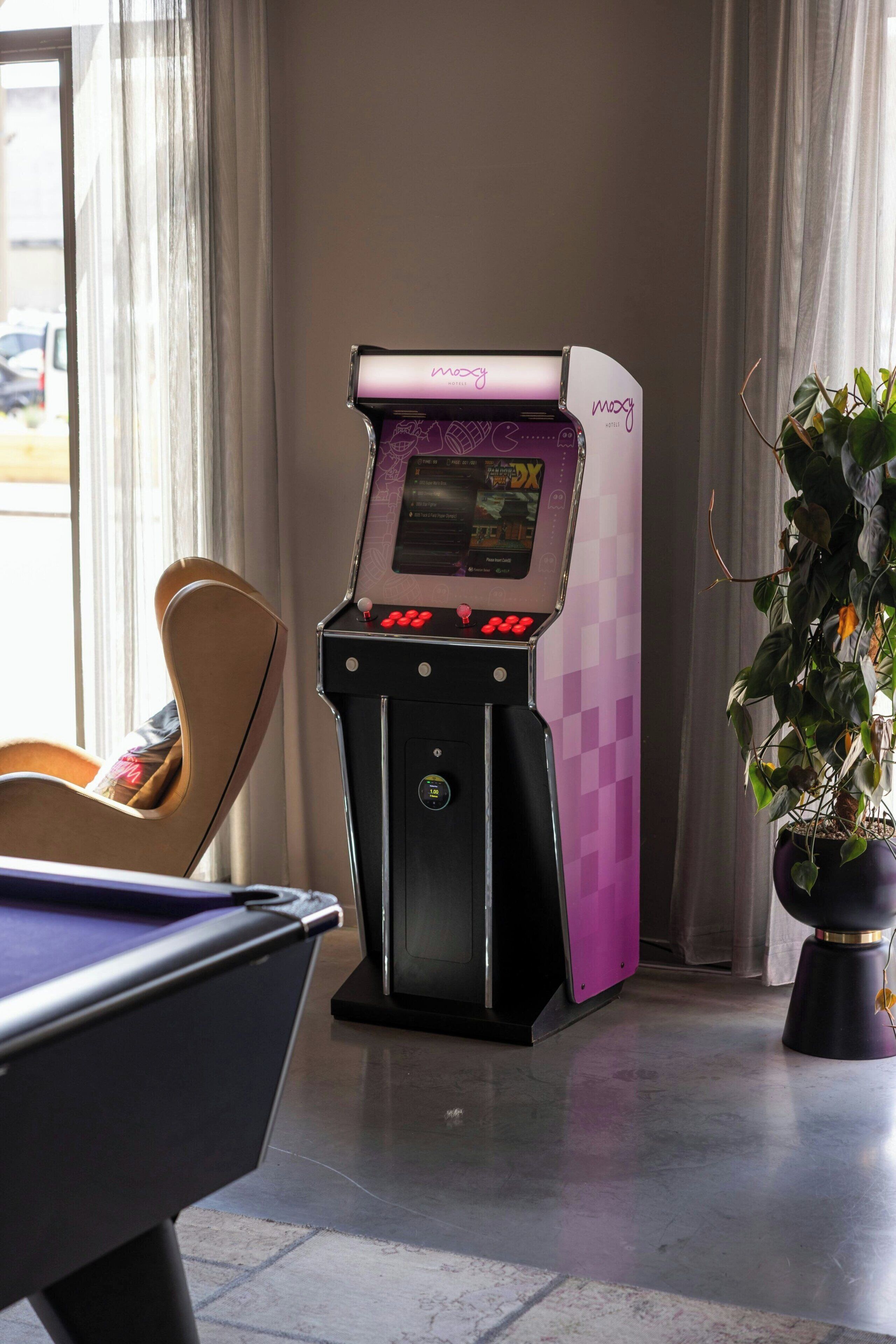 Una mesa de billar, una máquina de videojuegos púrpura y negra, una planta en maceta y una silla con cojín.