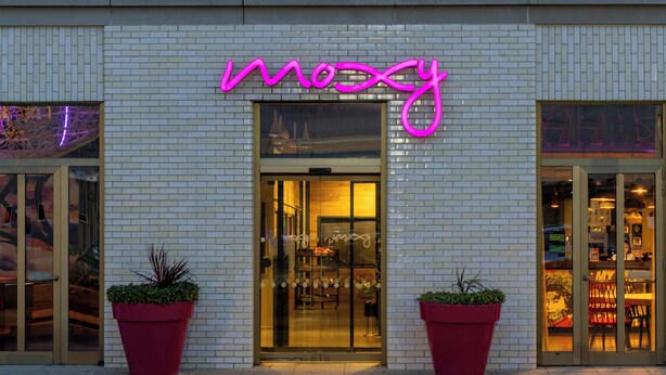 A building with a neon sign reading "moosy" above the entrance. There are two large potted plants in front of the entrance.