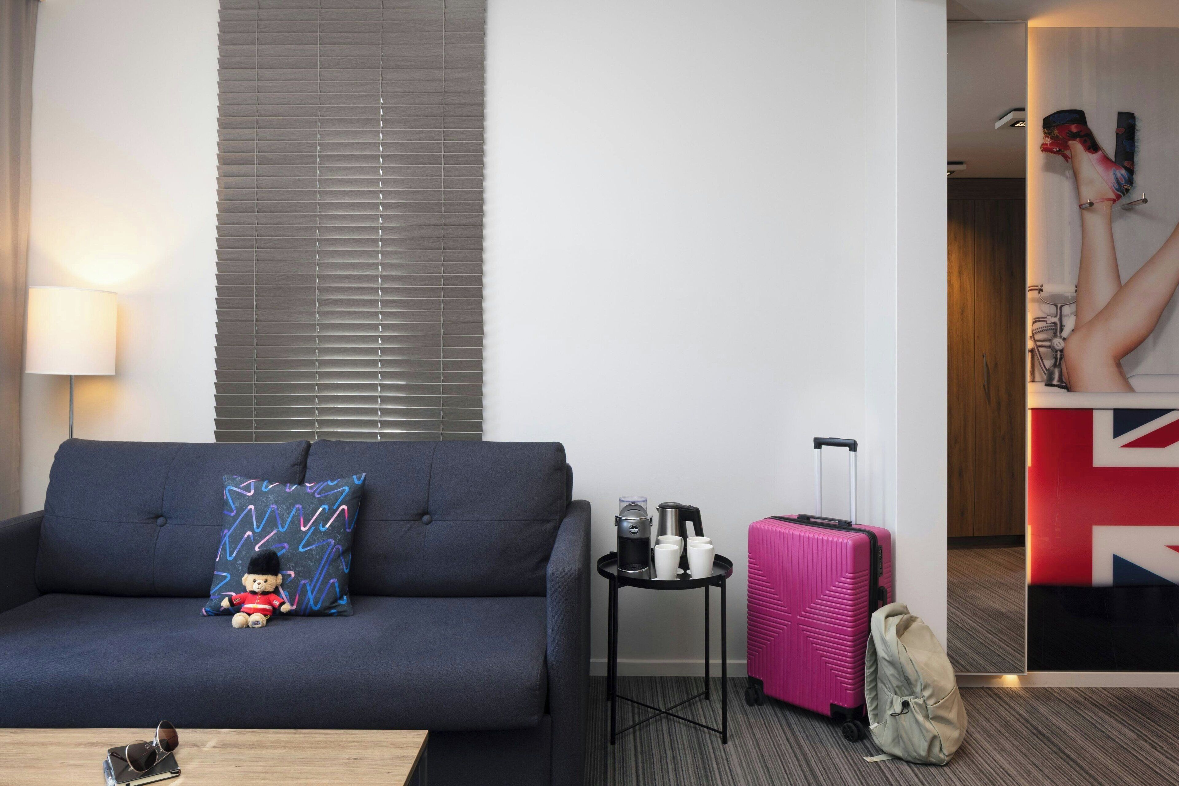 A modern hotel room with a dark blue sofa, a small table with a lamp, a pink suitcase, and a Union Jack flag on the wall.