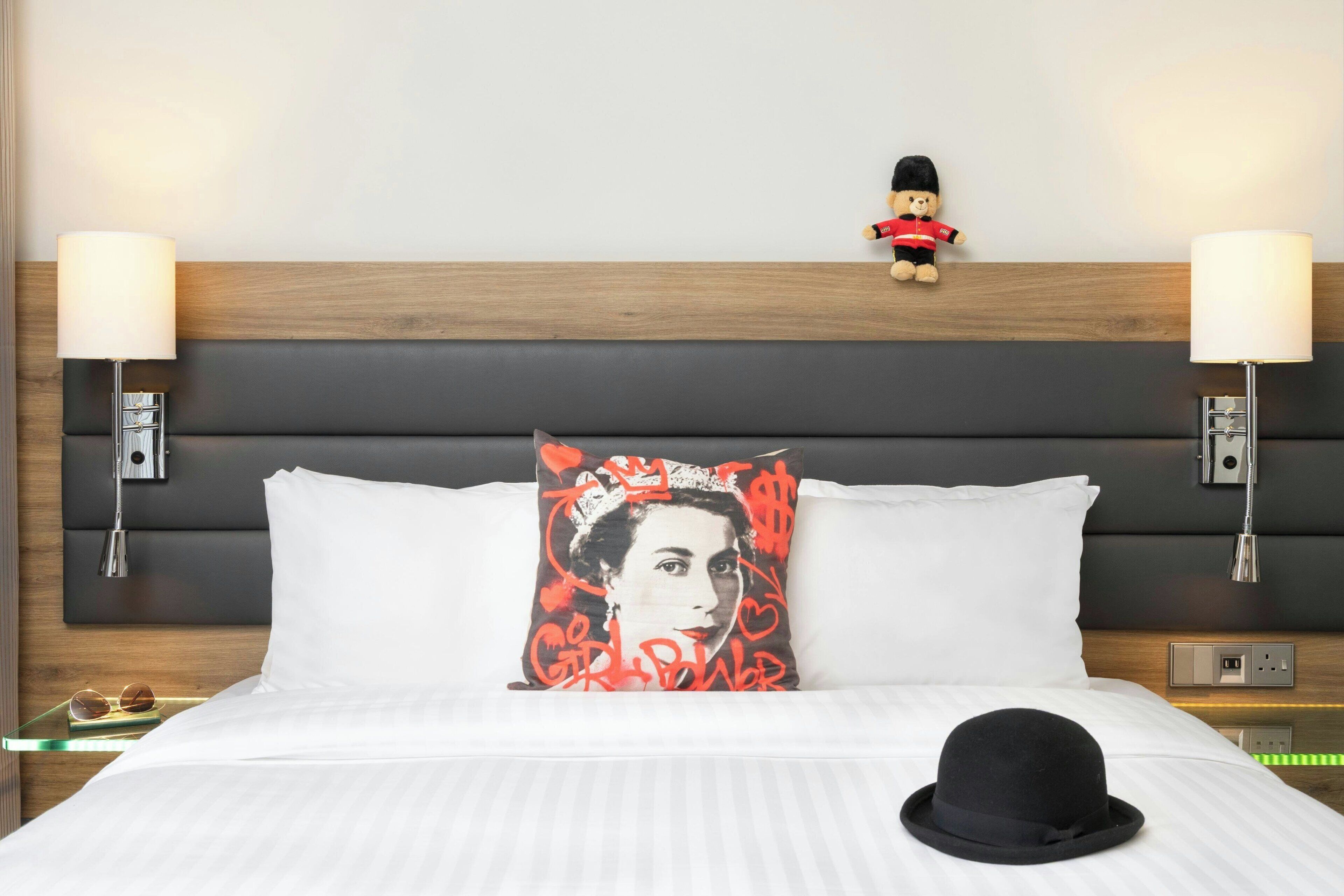 A hotel room with a bed, two bedside lamps, a nightstand, a decorative pillow, a teddy bear, and a black hat on the bed.