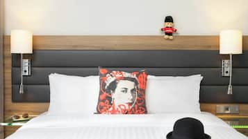 A hotel room with a bed, two bedside lamps, a nightstand, a decorative pillow, a teddy bear, and a black hat on the bed.