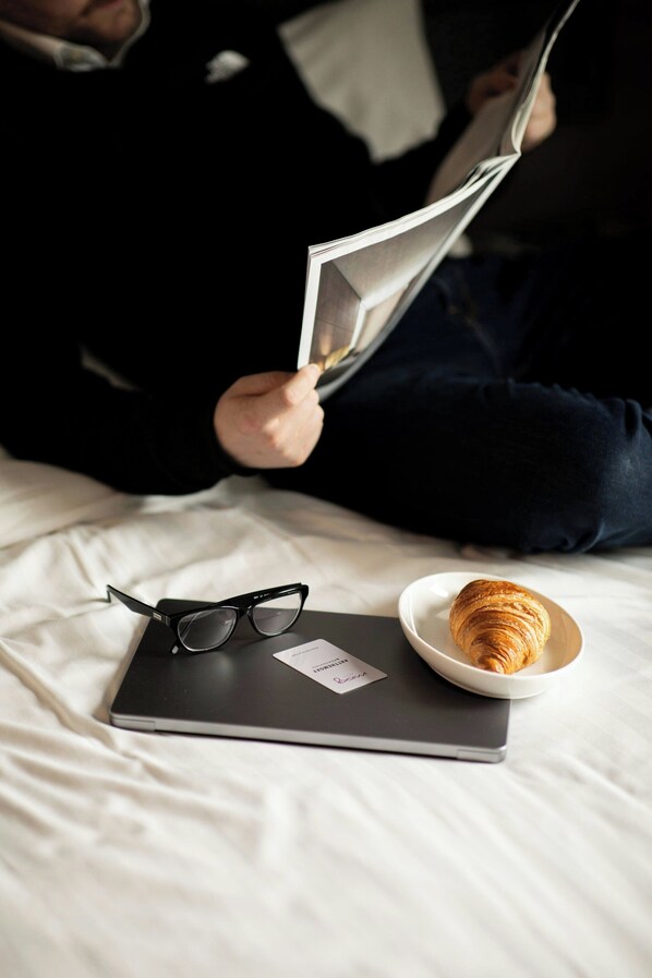 A laptop on a bed with a pair of glasses and a croissant. - Moxy London Excel (London)