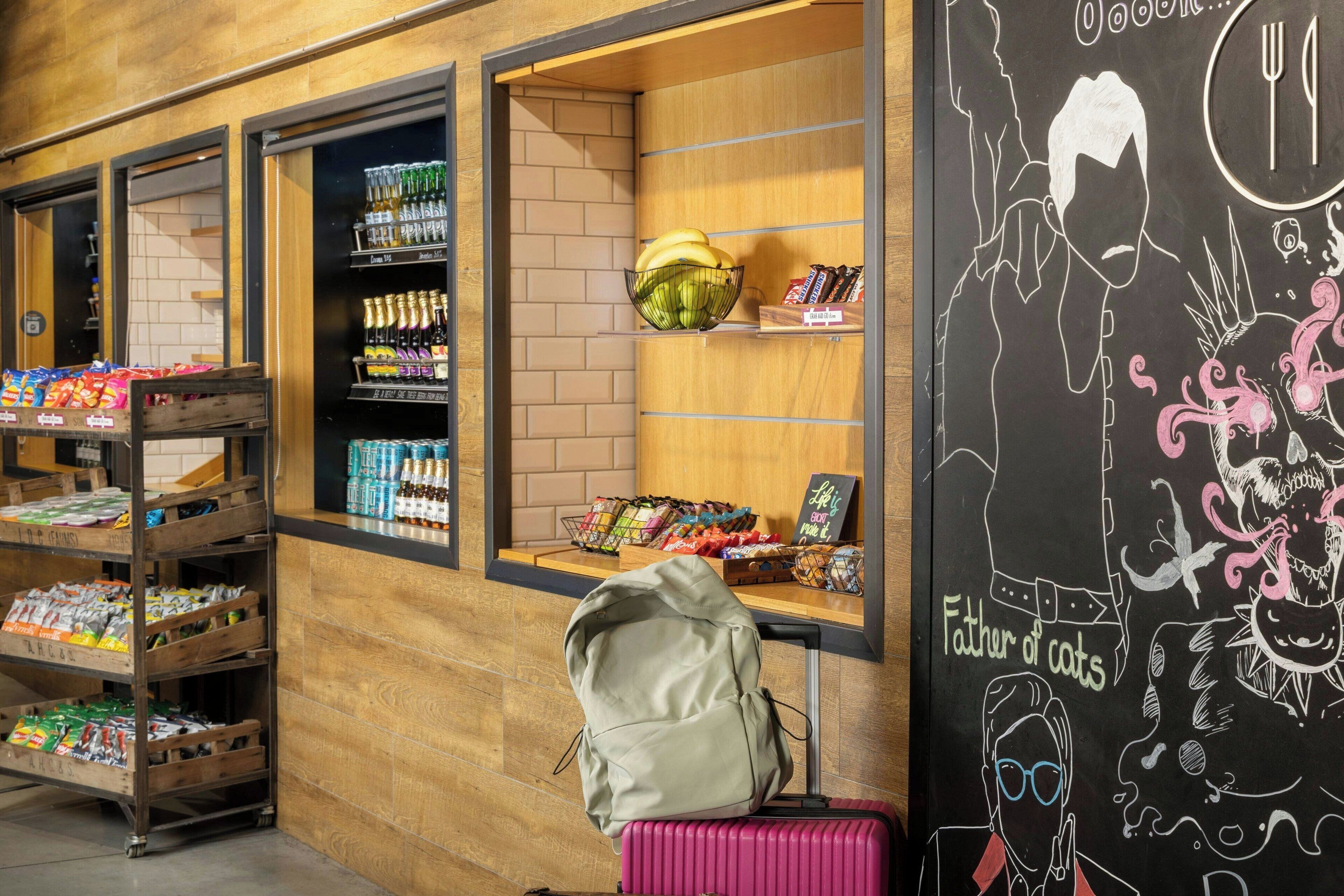 A store interior with shelves stocked with snacks, a display of bottled beverages, and a chalkboard with a cat illustration.