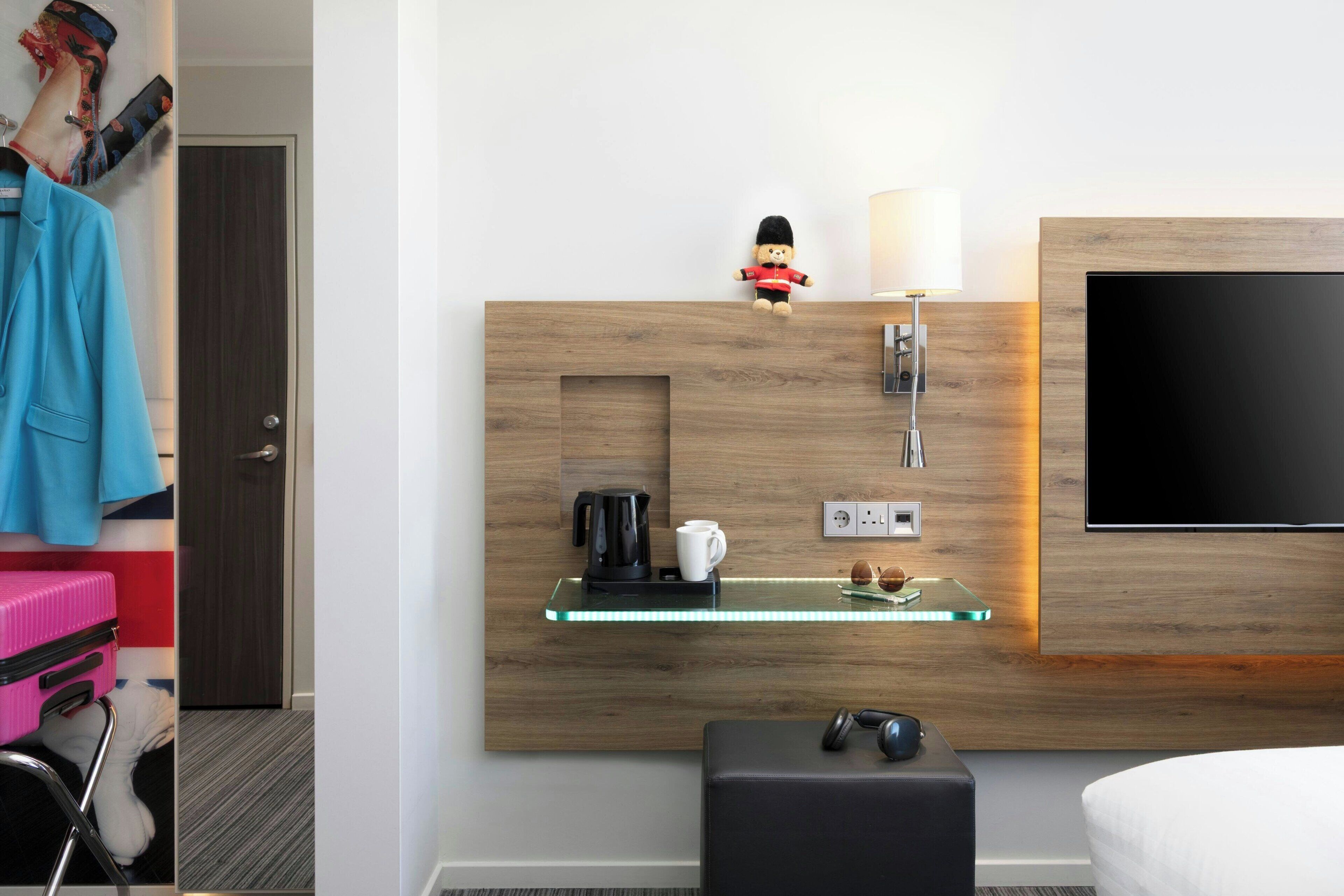 A hotel room with a wooden headboard, a flat-screen TV, a glass shelf with a coffee maker, a lamp, and a small bear figurine.