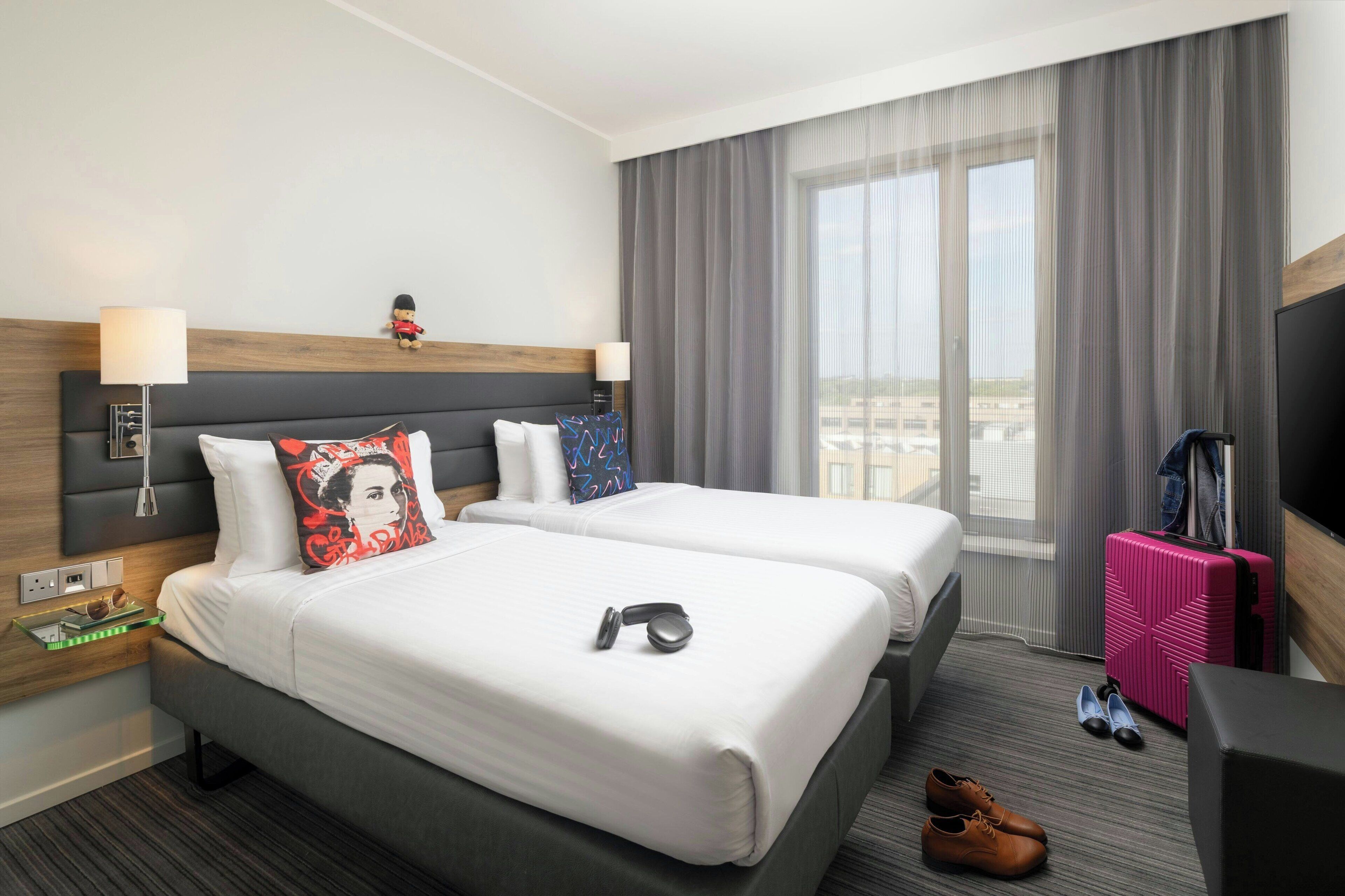 A hotel room with two beds, a TV, a desk, and a window with curtains.