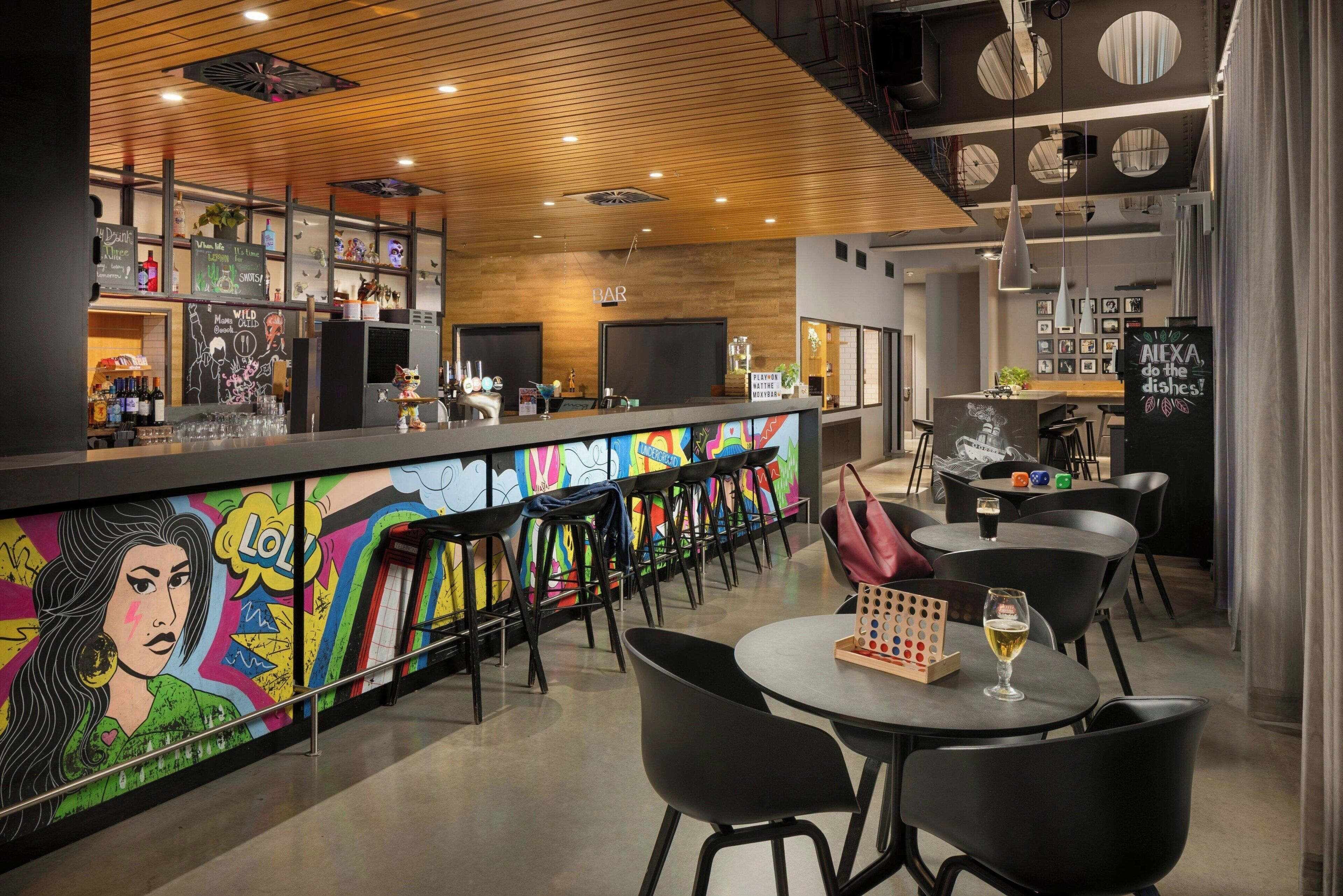 A modern bar area with a colorful mural, bar stools, and a round table with a glass of wine.