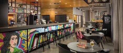 A modern bar area with a colorful mural, bar stools, and a round table with a glass of wine.