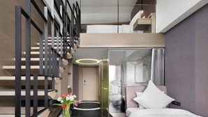 Une chambre d’hôtel moderne, équipée d’un lit, d’une petite table agrémentée de fleurs, d’une salle de bain fermée par des parois de verre et d’un escalier.