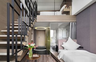 Una habitación de hotel moderna con cama, una mesita con flores, un baño con paredes de vidrio y una escalera.