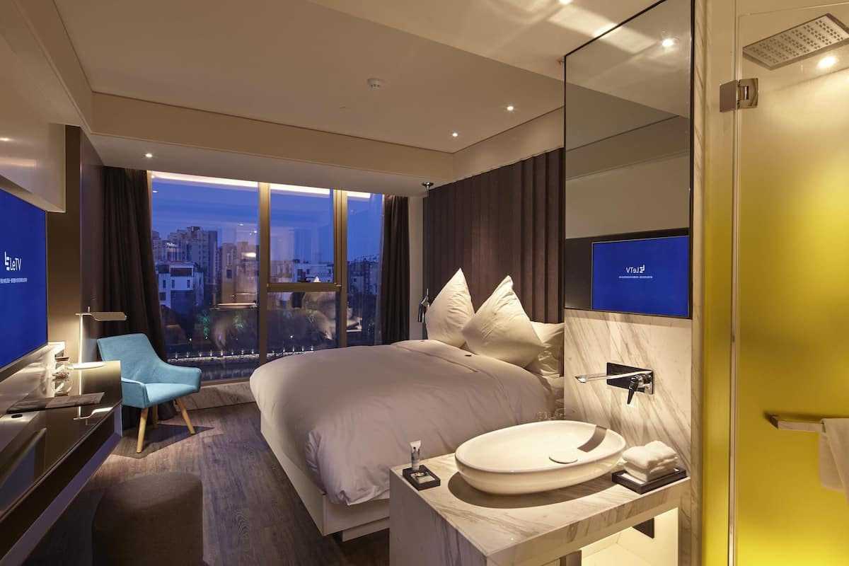 Ein modernes Hotelzimmer mit einem großen Bett, einem Schreibtisch, einem Stuhl, einem Badezimmer mit Waschbecken und Spiegel sowie Stadtblick durch große Fenster.