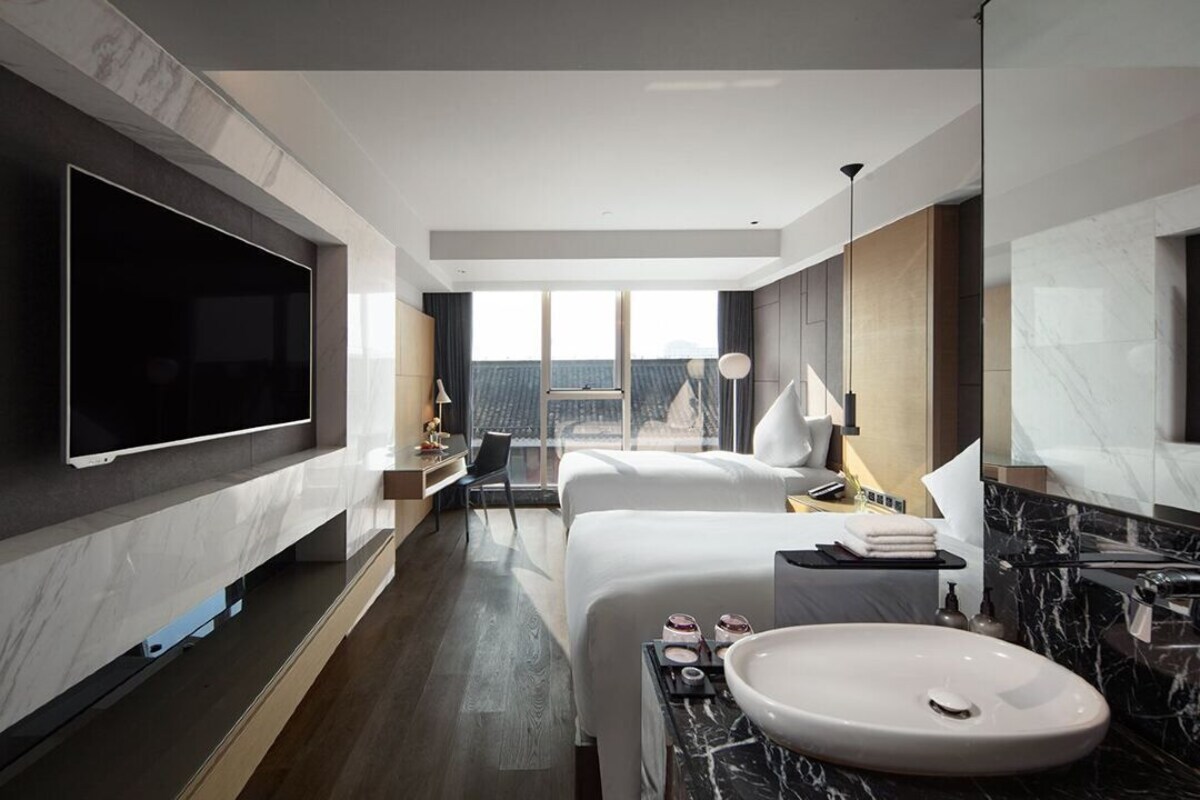 A modern hotel room with a large flat-screen TV, a bed, a bathroom with a sink and mirror, and a view of the city.
