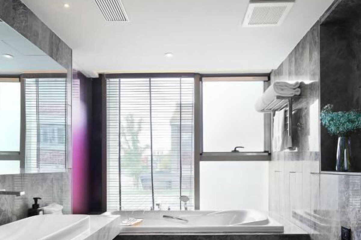 A modern bathroom with a large window, a bathtub, a toilet, and a vanity with a mirror.