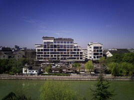 Un hotel con diseño arquitectónico tradicional y moderno, situado cerca de una masa de agua con paseo marítimo y zonas verdes.