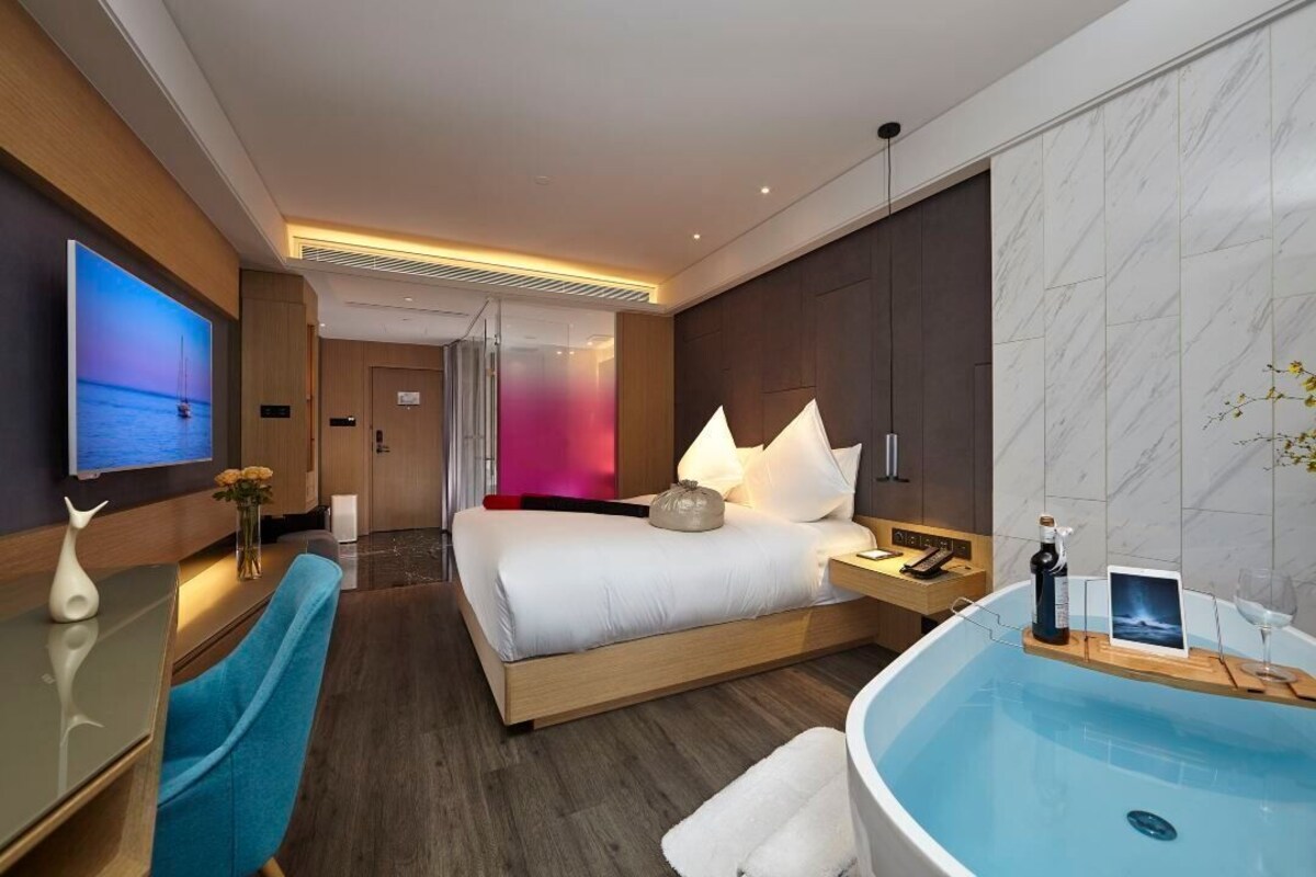 A modern hotel room with a large bed, a sitting area with a desk, a TV, a bathtub, and a view of the sea.