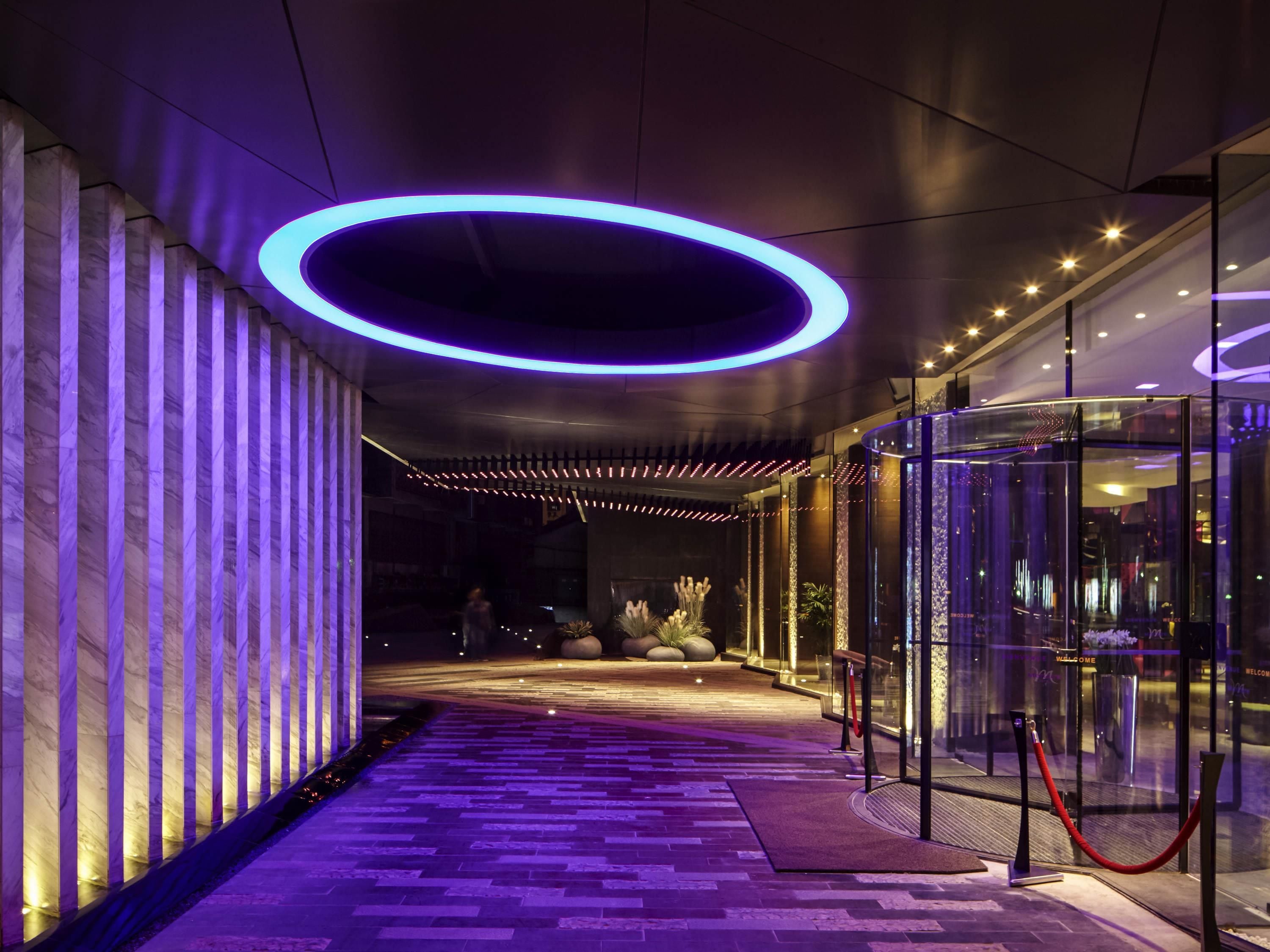 Ein moderner Hotelzugang mit einer Glastür, die sich dreht, einem roten Teppich und einem violett beleuchteten Weg.