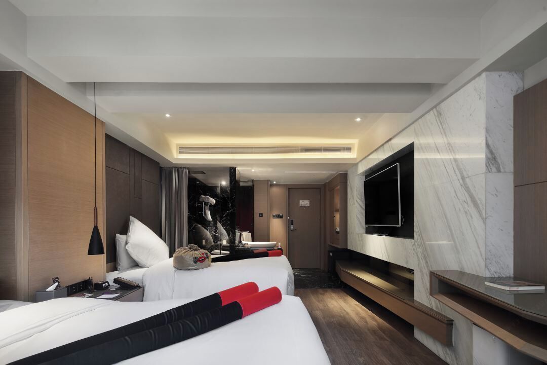 Una habitación de hotel moderna con un televisor de pantalla plana grande, una cama con ropa blanca y detalles rojos, una mesita de noche con lámpara y una pared de acento de mármol.