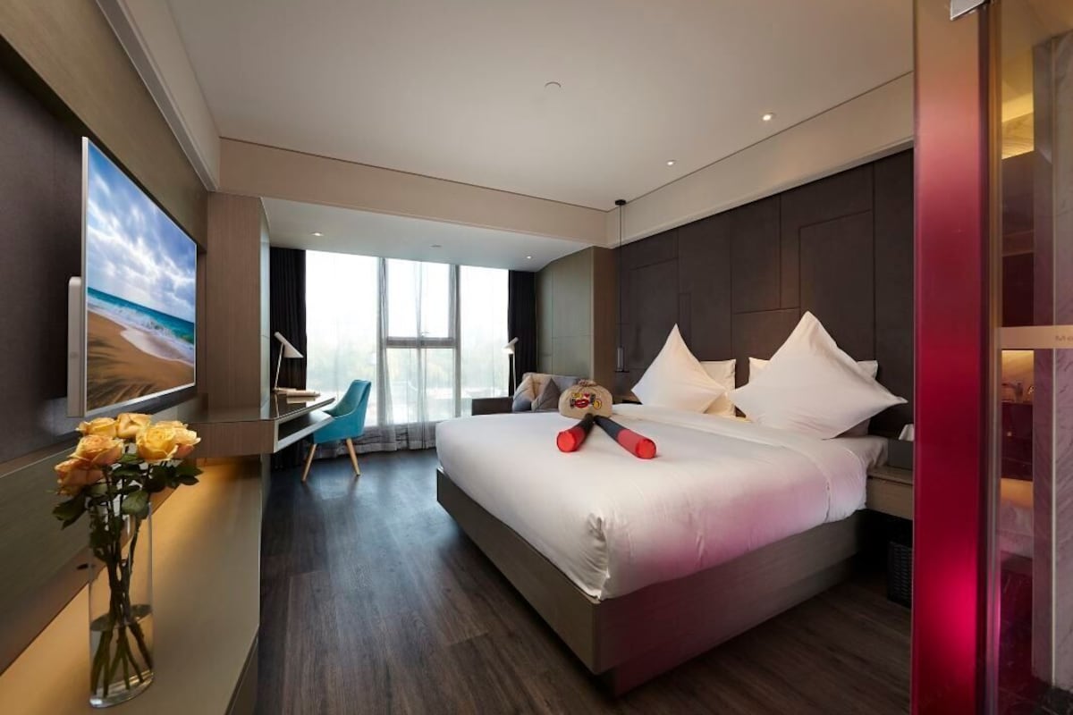 A modern hotel room with a large bed, a desk, a chair, and a large window.