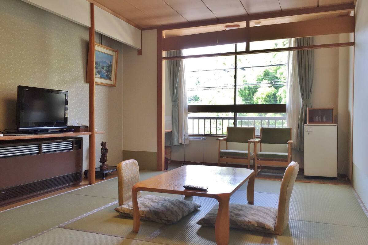 Quarto tradicional japonês com piso de tatame, uma mesa baixa e cadeiras. Uma televisão está em um armário e há uma janela com vista para o verde lá fora.