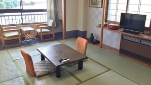 Una habitación tradicional japonesa con suelo de tatami, una mesa baja con cojines, un televisor y una ventana con vistas a la vegetación.