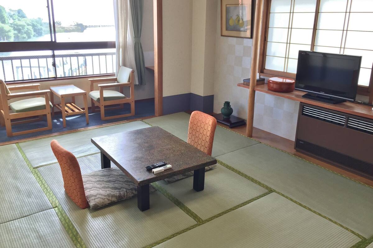 Um quarto japonês tradicional com tatames, uma mesa baixa com almofadas, uma televisão e uma janela com vista para o verde.