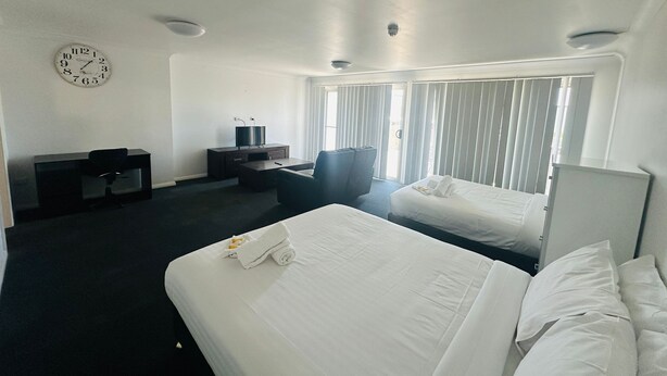 A hotel room with two beds, a desk with a computer, a chair, a clock on the wall, and large windows with blinds. - Aspire Mayfield (Mayfield)