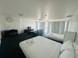 Habitación de hotel con dos camas, un escritorio con computadora, una silla, un reloj en la pared y ventanas amplias con persianas.