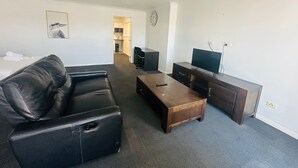 A living room with a leather sofa, a wooden coffee table, a TV on a wooden stand, and a kitchen in the background. - Aspire Mayfield (Mayfield)