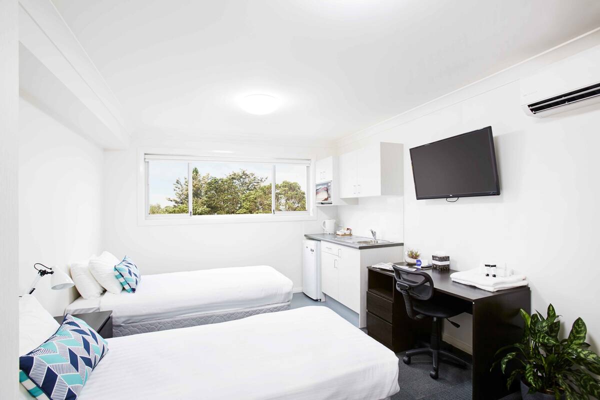 A hotel room with two beds, a desk, a TV, a window, and a small kitchenette.