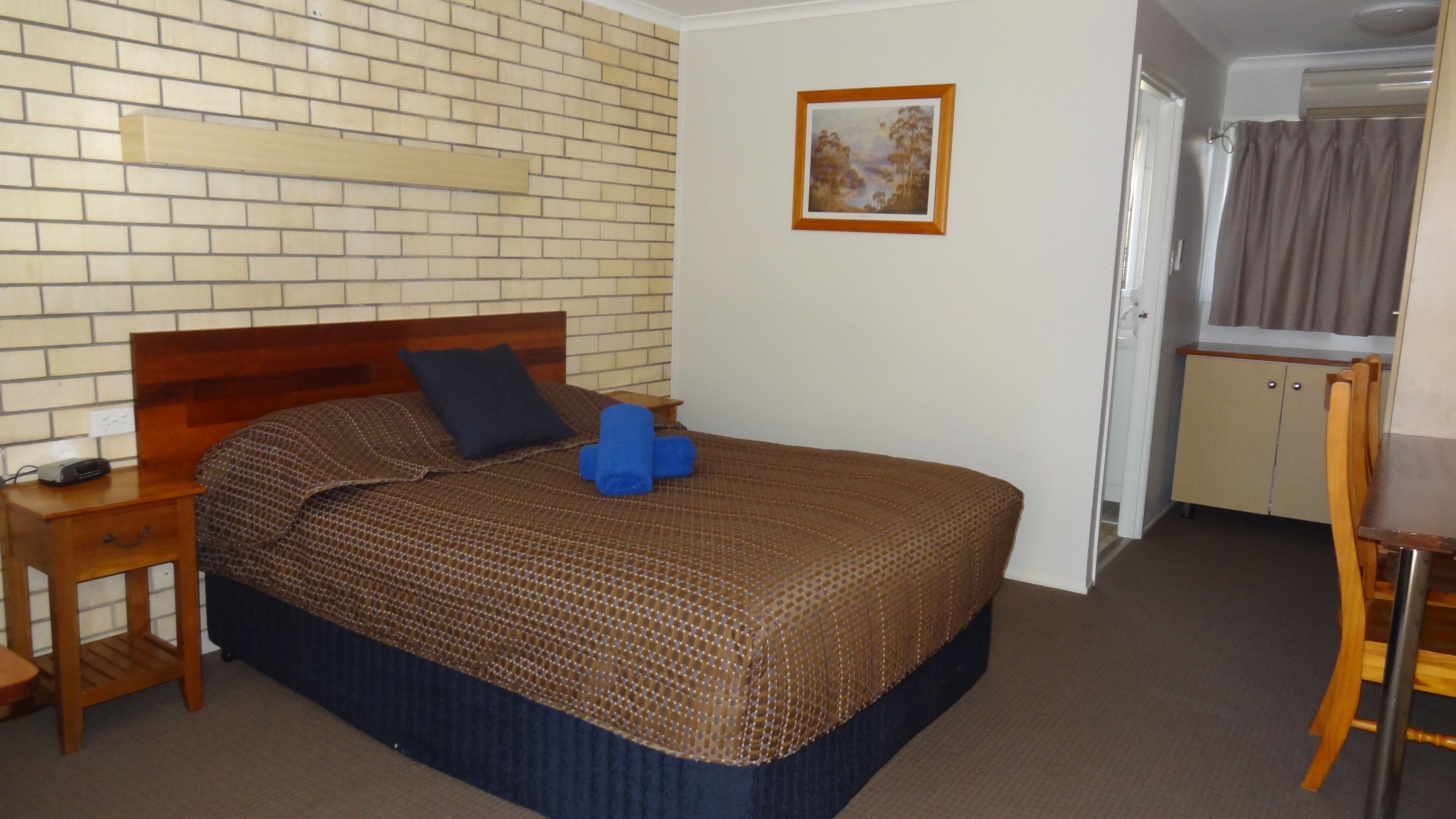 A bedroom with a brick-patterned wall, a wooden headboard, a bed with a brown patterned bedspread, two blue pillows, a bedside table with a lamp, and a small wooden chair.