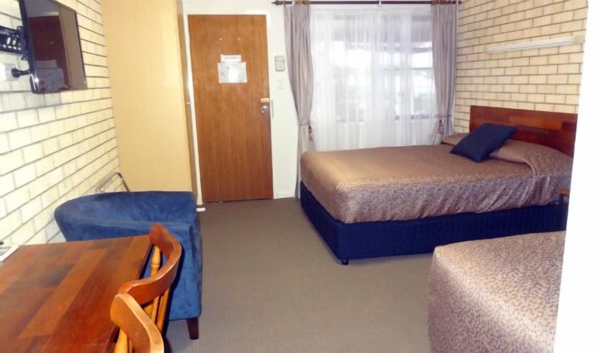 A small, single-room hotel room with a wooden door, a bed with a blue headboard, a small table with a chair, and a window with curtains.