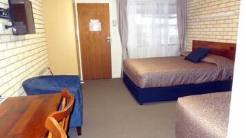A small, single-room hotel room with a wooden door, a bed with a blue headboard, a small table with a chair, and a window with curtains.