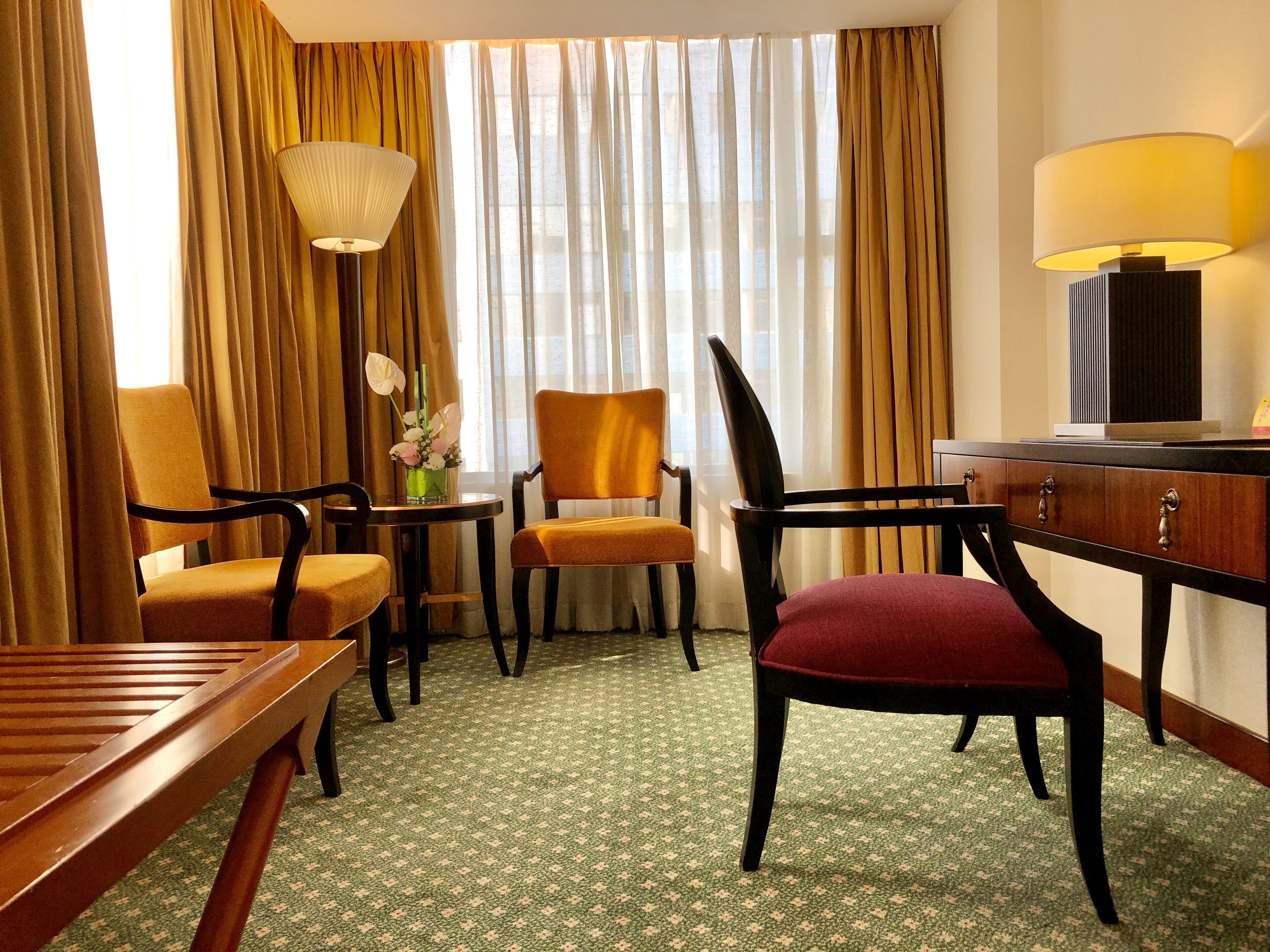 Una habitación de hotel con un escritorio de madera, dos sillas, una mesita con un florero y una lámpara.