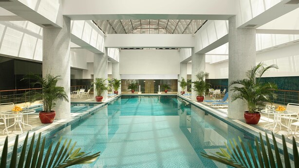 Una amplia piscina interior con paredes y techo azulejados, rodeada de sillones de descanso y plantas en macetas.