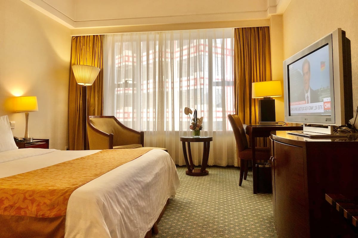 Habitación de hotel con una cama, un escritorio con computadora, una silla, una pequeña mesa con un florero y una ventana con cortinas.