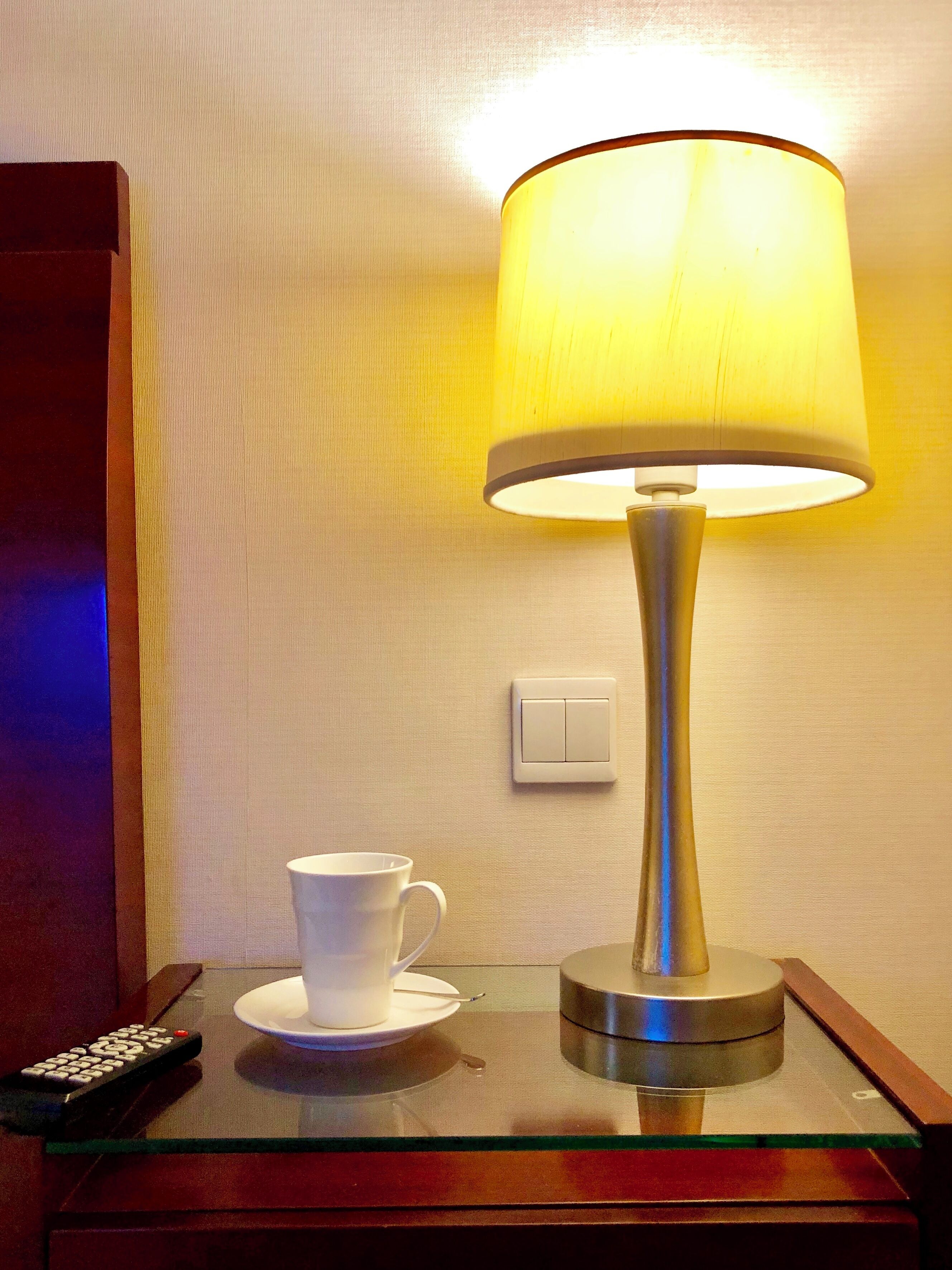 A table with a white cup and saucer, a remote control, and a lamp with a yellow shade.