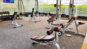 Un gimnasio con diverso equipo de ejercicio, un televisor y amplias ventanas con vista a la vegetación.